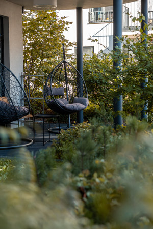 Outdoor patio area features two modern hanging egg chairs with cushions, surrounded by dense greenery. A building with balconies is visible in the background.の写真素材