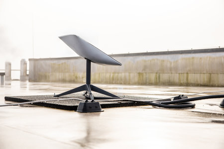 A satellite dish is mounted on a wet rooftop, with cables extending from its base. The clear sky and weathered wall suggest a modern urban setting.の写真素材