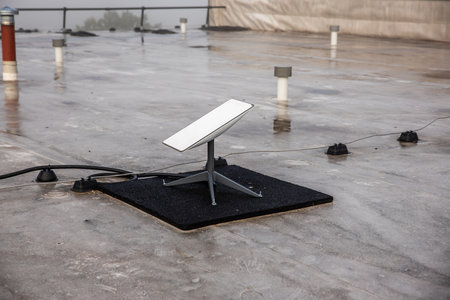 A rooftop features a satellite dish on a wet surface, with small vents and pipes. An overcast sky and railing suggest an urban setting.の写真素材