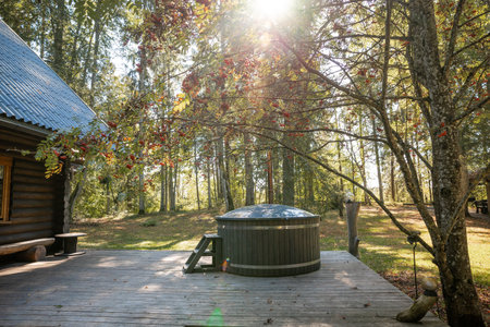 A wooden deck features a round hot tub surrounded by lush greenery and tall trees. Sunlight filters through branches, with a rustic cabin partially visible.の写真素材