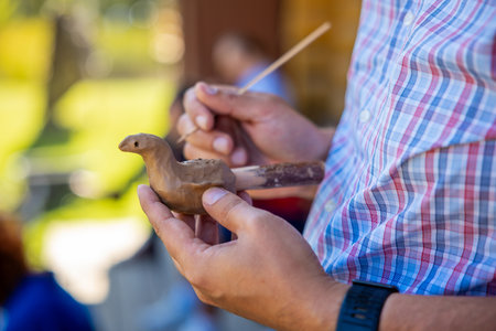 A person in a plaid shirt holds a clay bird sculpture and a wooden tool, focusing on the art of crafting in a blurred outdoor setting with greenery.の写真素材