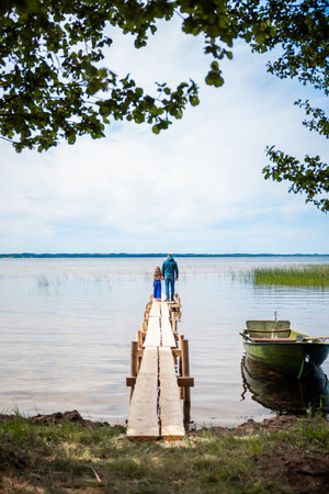 A father and daughter stand on a wooden boardwalk on the shore of a lake and look into the waterの写真素材