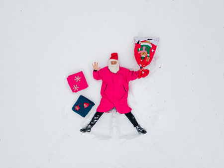 joyful santa claus in a vibrant pink suit lying in the snow, surrounded by colorful christmas gifts and a festive stocking, celebrating the winter season's happiness and holiday spiritの写真素材