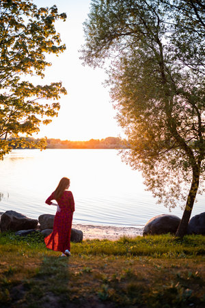 woman in red dress enjoying a peaceful sunset by the lakeside, the golden sunlight casting a warm glow on the serene watersの写真素材