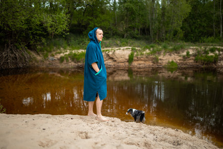 Man in Teal Towel Poncho poses by River, Watchful Dog Stands Nearbyの写真素材