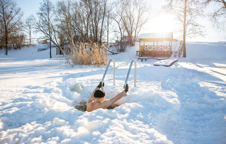 Young man enjoying winter swimming in a hand-cut ice hole on a sunny snowy dayの写真素材
