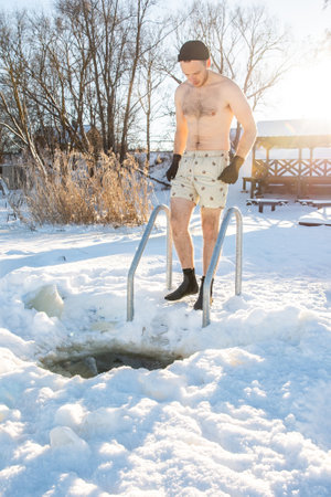 Confident man emerging from an ice swimming hole in winter, clad in swim shorts against snowy backdropの写真素材