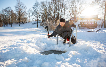 Man carving out a hole in the ice with an axe for winter swimming in cold sunny winter dayの写真素材