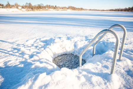Beautiful inter landscape with a hole cut in the snow-covered ice for cold water swimming, complete with a frosted metal ladder, under a clear blue skyの写真素材