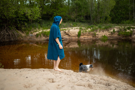 Man in Teal Poncho Standing by the River, Dog Playing by the Riversideの写真素材