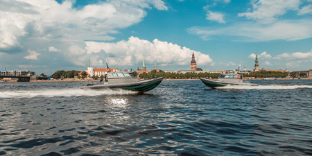 Two military speed boats patrolling the river with a historic city skyline and fluffy white clouds in the backgroundの写真素材
