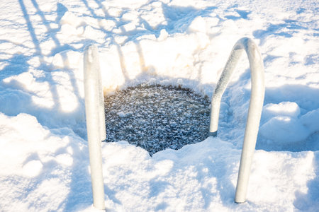 Bright winter day with a frosted ladder leading into a natural ice swimming hole on a snow-covered lakeの写真素材