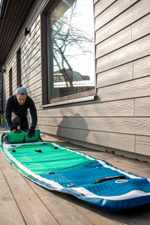 Man folding an inflatable stand-up paddleboard (SUP) on a wooden deck next to a modern home
