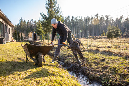 Man in protective gear tirelessly working to dredge a muddy trench with a shovel and wheelbarrow in a backyard during early spring.の写真素材