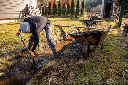 Man in protective gear tirelessly working to dredge a muddy trench with a shovel and wheelbarrow in a backyard during early spring.の写真素材