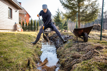 Man in workwear clearing a waterlogged garden path in early spring with a shovel and wheelbarrow amidst a rural landscape.の写真素材