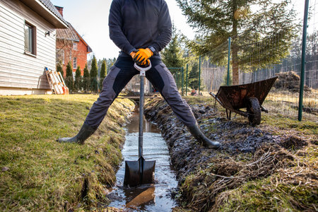 Man in protective gear tirelessly working to dredge a muddy trench with a shovel and wheelbarrow in a backyard during early spring.の写真素材