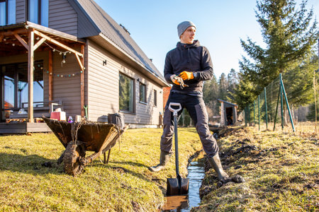 Man in protective gear tirelessly working to dredge a muddy trench with a shovel and wheelbarrow in a backyard during early spring.の写真素材