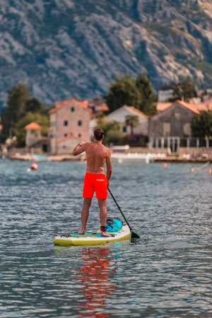 man enjoying stand-up paddleboarding on serene blue waters with a picturesque coastal town backdrop on a sunny dayの写真素材