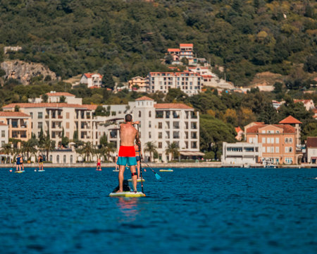man enjoying stand-up paddleboarding on serene blue waters with a picturesque coastal town backdrop on a sunny dayの写真素材