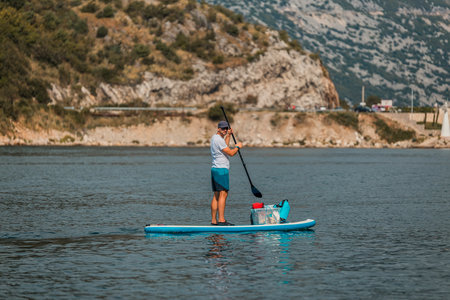 man enjoying stand-up paddleboarding on a calm sea with mountainous landscape in the backgroundの写真素材