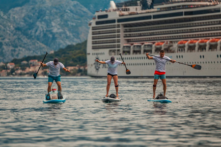 three paddle boarders on a scenic adventure in front of a majestic cruise ship with mountain backdropの写真素材