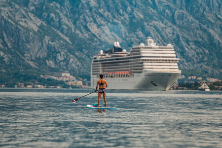 solo paddle boarder in front of a towering cruise ship with a stunning mountainous backdropの写真素材