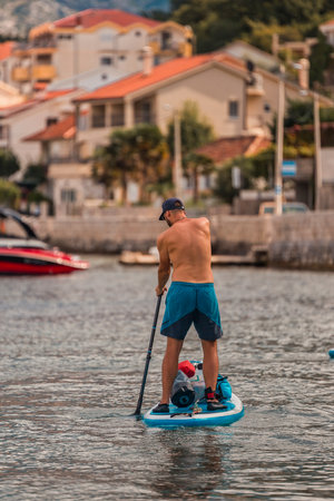 man enjoying stand-up paddleboarding on serene blue waters with a picturesque coastal town backdrop on a sunny dayの写真素材