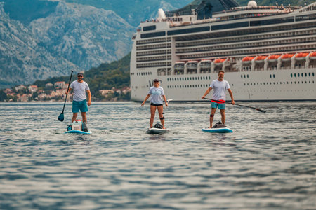 three paddle boarders on a scenic adventure in front of a majestic cruise ship with mountain backdropの写真素材