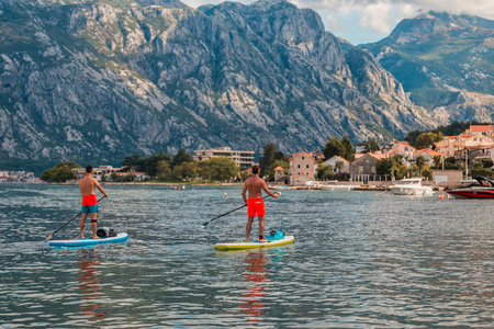 man enjoying stand-up paddleboarding on serene blue waters with a picturesque coastal town backdrop on a sunny dayの写真素材