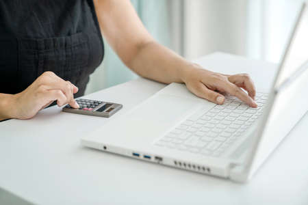 Woman is sitting at laptop and doing calculations on calculator. Hands, calculator, and laptop...