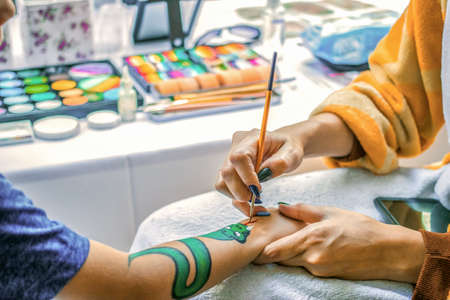 Young girl artist draws a dragon on childs hand. Painting skin with paints, close-up, without faces.