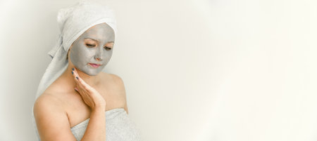 Young woman with clay cosmetic mask, wrapped in towel, touch face with hand, her gaze is directed downward. Facial skin care with cosmetic treatment. Copy space, bannerの写真素材