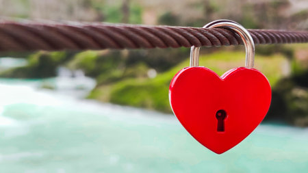 Castle in shape of red heart hangs on metal cable against background of river and nature. Concept of...