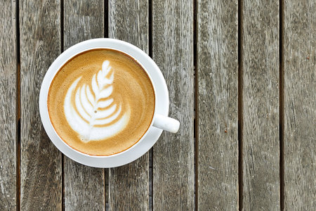 White cup of coffee with pattern on top of foam, against background of wooden table, copy space, top...