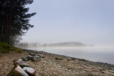 Lakeside shore and islands hidden behind thick fog. Stones on the coast; sand and pebble beach; pines silhouettes. Early morning; misty landscape.の写真素材