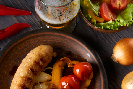 Chicken sausage with vegetables and light beer on a clay plate. Tomatoes, lettuce, sweet peppers, onions, rye bread. Close-up, top view, dark wooden background.の写真素材