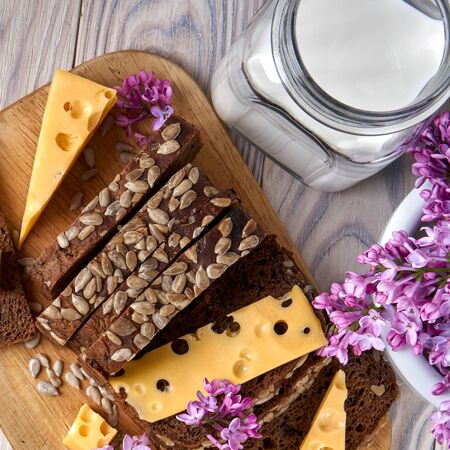 Dark multigrain bread made from rye, wheat, oats and barley with sunflower and flax seeds. Healthy natural nutrition. Fresh farm food. Milk, cheese, lilac flowers, rustic bleached wood table.の写真素材