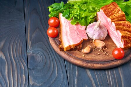 Piece of smoked pork belly with sliced strip and grain bread on a wooden cutting board. Vegetables and seasonings: tomatoes, lettuce, parsley, garlic and coriander. Rustic style.の写真素材
