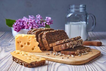 Dark multigrain bread made from rye, wheat, oats and barley with sunflower and flax seeds. Healthy natural nutrition. Fresh farm food. Milk, cheese, lilac flowers, rustic bleached wood table.の写真素材