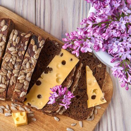 Dark multigrain bread made from rye, wheat, oats and barley with sunflower and flax seeds. Healthy natural nutrition. Fresh farm food. Cheese, lilac flowers, rustic bleached wood table.の写真素材