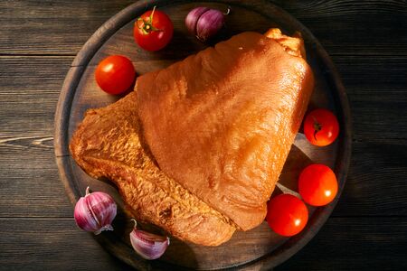 Smoked pork knuckle with fresh cherry tomatoes and garlic, illuminated by the slanting rays of the sun. Rustic cutting board and wooden background.の写真素材