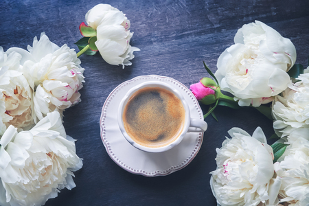 A cup of coffee and white peonies on dark wooden background. Flat-lay, top view, copy space. Toned imageの写真素材