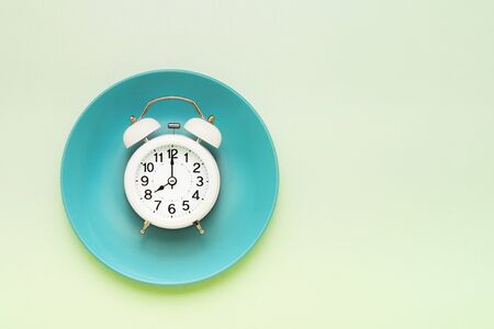 Intermittent fasting concept. White alarm clock on empty green plate on green background. Top view,...