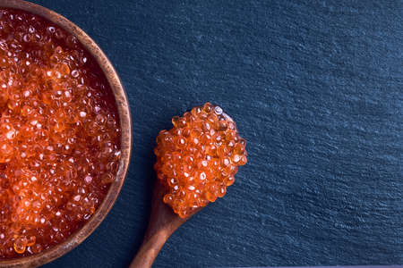 Red caviar of trout fish in a wooden bowl and spoon with caviar on a black slate background. Top view, copy space for textの写真素材
