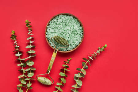 Green face roller from natural jade stone, bath salt and eucalyptus branches on a red background. SPA concept.の写真素材