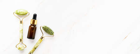 Green face rollers from natural jade stone and dark brown glass dropper bottle for medical and cosmetic use on a marble background. SPA concept. Top view, copy space, bannerの写真素材