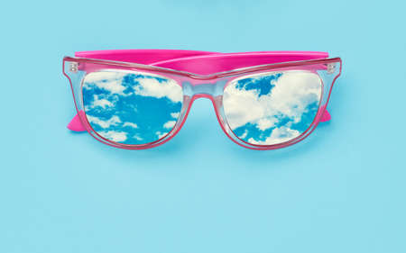 Pink sunglasses with cloudy sky reflection on blue background. Summer vacations concept. Copy spaceの写真素材