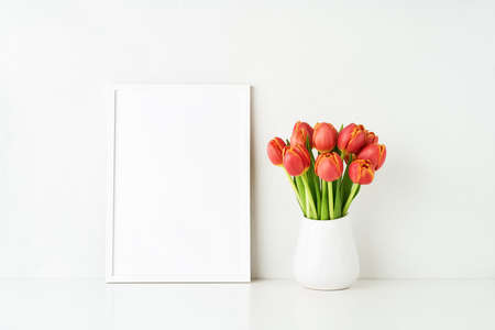 White frame mockup and red tulips in a white vase on a white table by the wall. Copy space