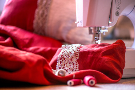 A macro shot of red and white thread spools and a white, crochet natural linen lace trim on a dark red linen fabric, being used while sewing tablecloth by using a sewing machineの写真素材